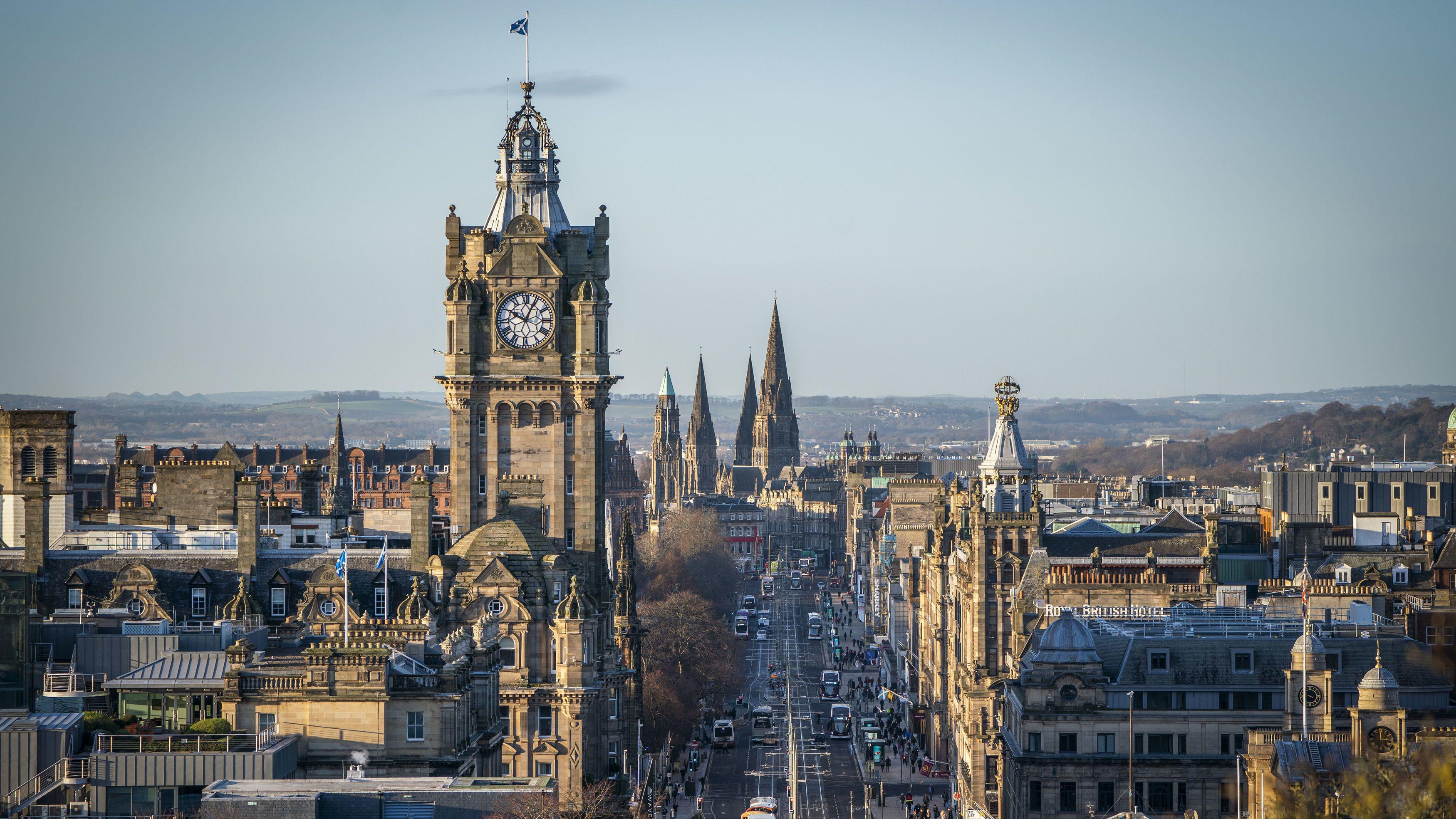 A imagem mostra uma vista panor&acirc;mica de Edimburgo, cidade marcada por edif&iacute;cios hist&oacute;ricos de pedra, com uma imponente torre de rel&oacute;gio dominando o horizonte. A avenida principal se estende em linha reta pelo centro da cena, cercada por constru&ccedil;&otilde;es de arquitetura vitoriana e neog&oacute;tica, enquanto torres e pin&aacute;culos se destacam ao fundo sob um c&eacute;u claro.
