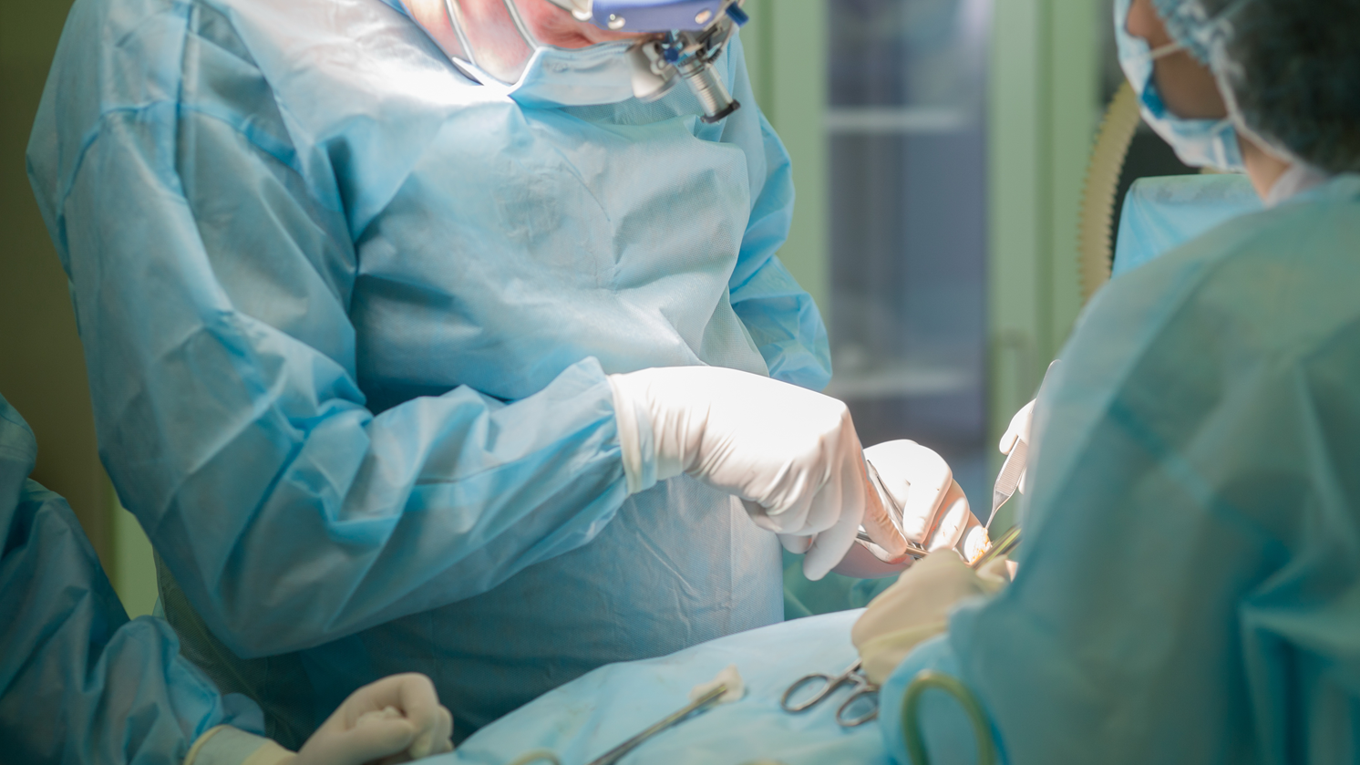 M&eacute;dicos vestidos de azul, com m&aacute;scaras e luvas brancas, realizando uma cirurgia