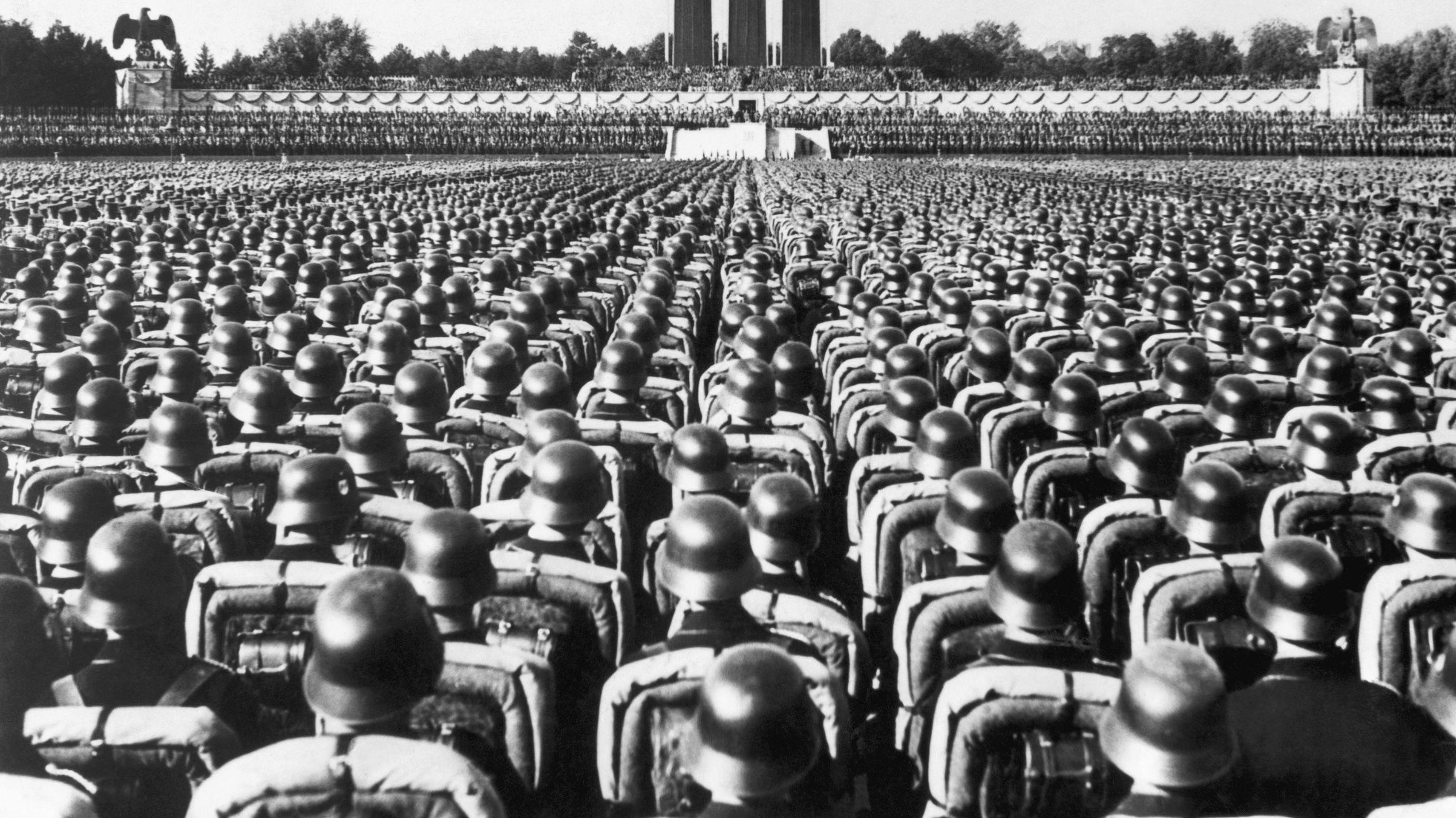 Foto em preto e branco mostra milhares de soldados com capacetes pretos, voltados para um grande palco com tr&ecirc;s grandes colunas. O Partido Nazista realizava com&iacute;cios semelhantes aqui todos os anos entre 1933 e 1938. 