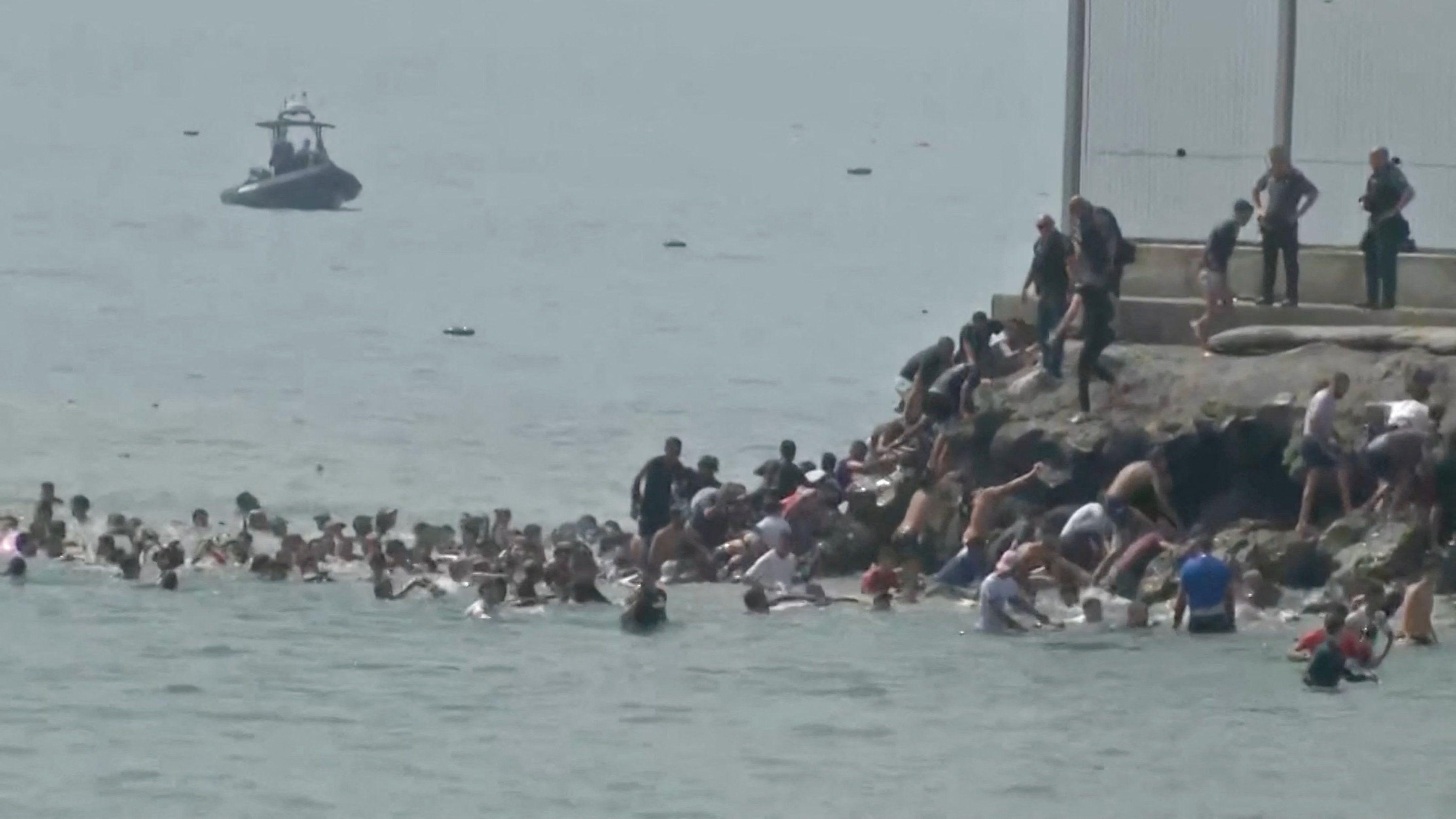 Decenas de personas permanecen de pie en el mar mientras otras suben por una pequeña orilla para desembarcar. Agentes con uniformes de seguridad vigilan desde la orilla. Alguien extiende la mano mientras se dirige hacia las personas que suben por la orilla. Al fondo, se ve una pequeña embarcación en el mar.
