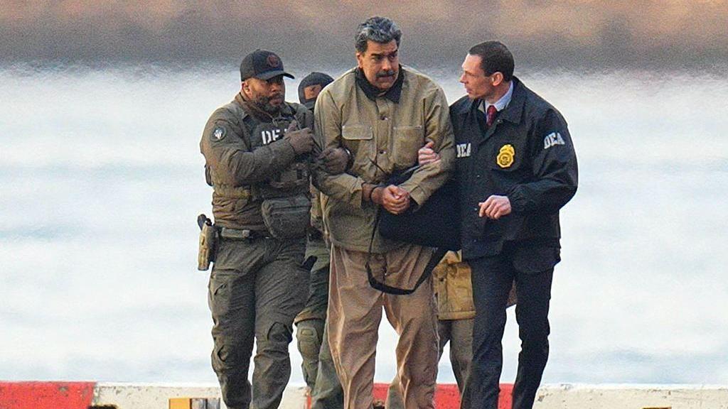 Nicolás Maduro in handcuffs wearing a beige coat and trousers after landing at a Manhattan helipad. Two men hold his arms and two more people follow behind.