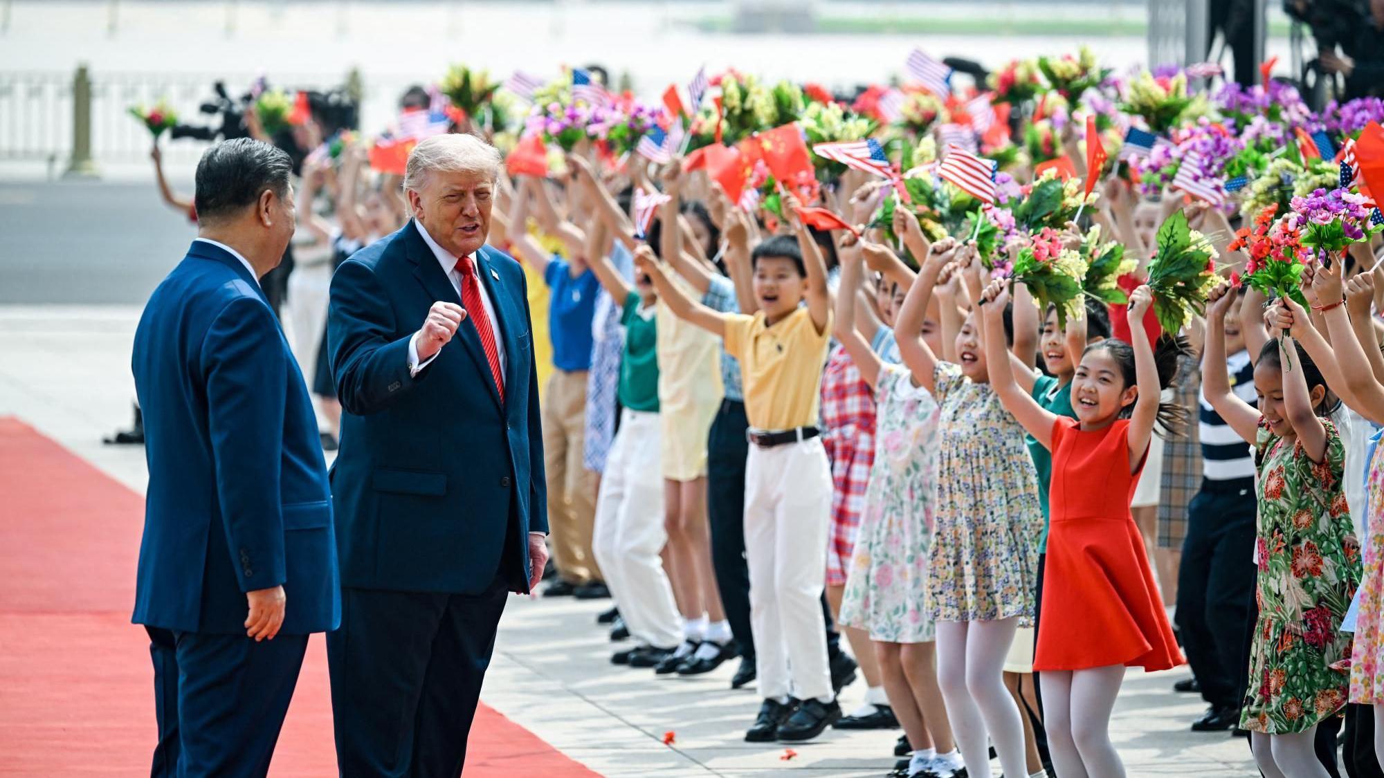 El presidente Trump se sitúa junto al presidente Xi con el puño en alto, mientras ambos caminan por una alfombra roja flanqueada por filas de niños que vitorean y ondean flores, así como banderas chinas y estadounidenses.