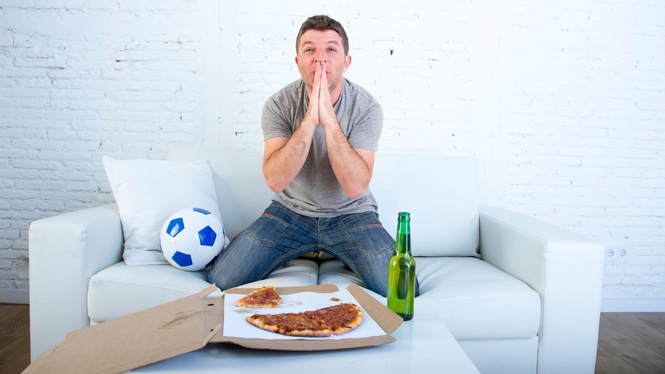 شاب يجلس على مقعد في وضعية مشاهدة مباراة على شاشة وأمامه قطعة بيتزا وزجاجة شراب وبجانبة كرة