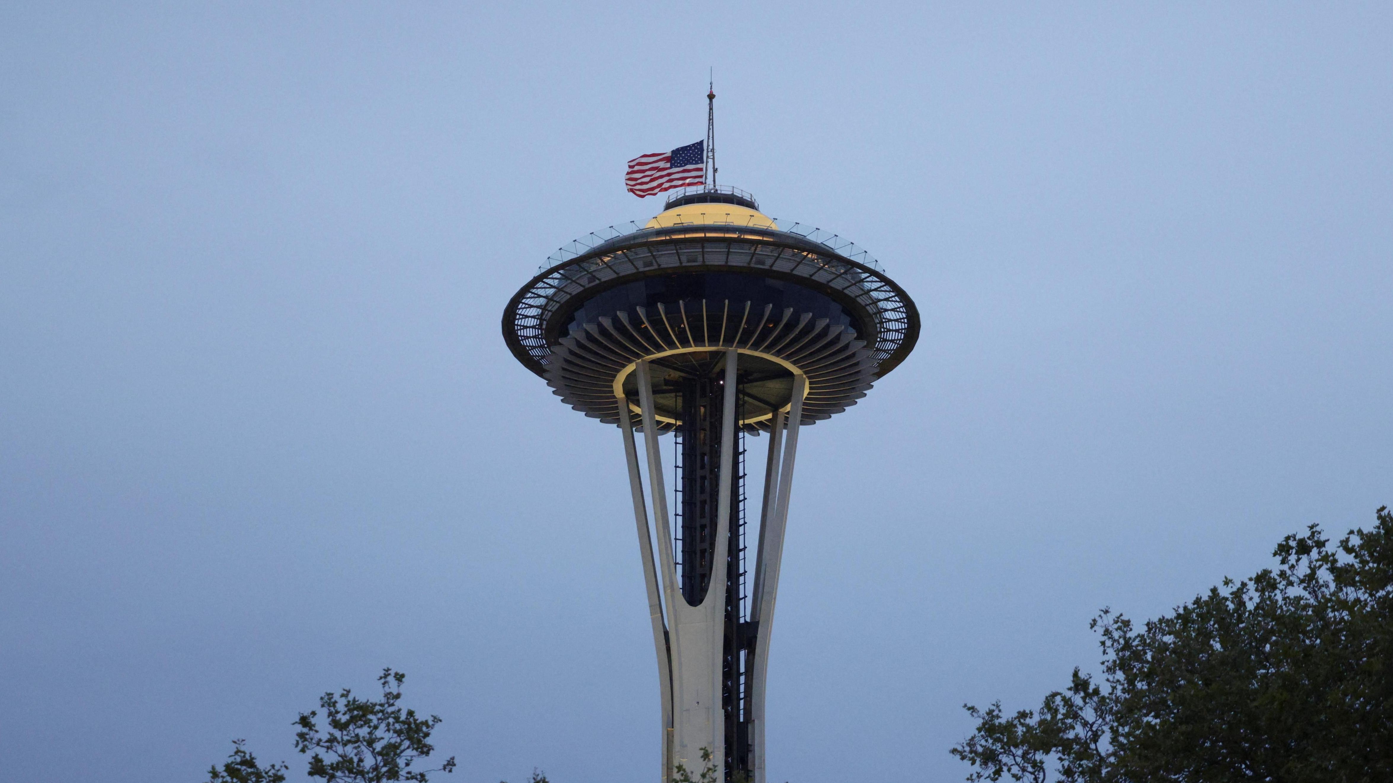 O edif&iacute;cio Space Needle de Seattle ao fundo, ao entardecer. Uma grande bandeira dos EUA est&aacute; hasteada a meio mastro em homenagem &agrave;s v&iacute;timas do tiroteio.