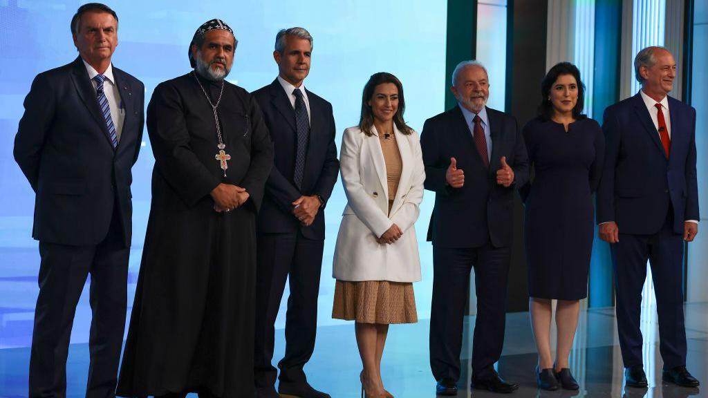 Foto em um estúdio de TV com Bolsonaro, padre Kelmon, Felipe d'Avila, Soraya Thronicke, Lula, Simone Tebet e Ciro Gomes, da esquerda para a direita.  
