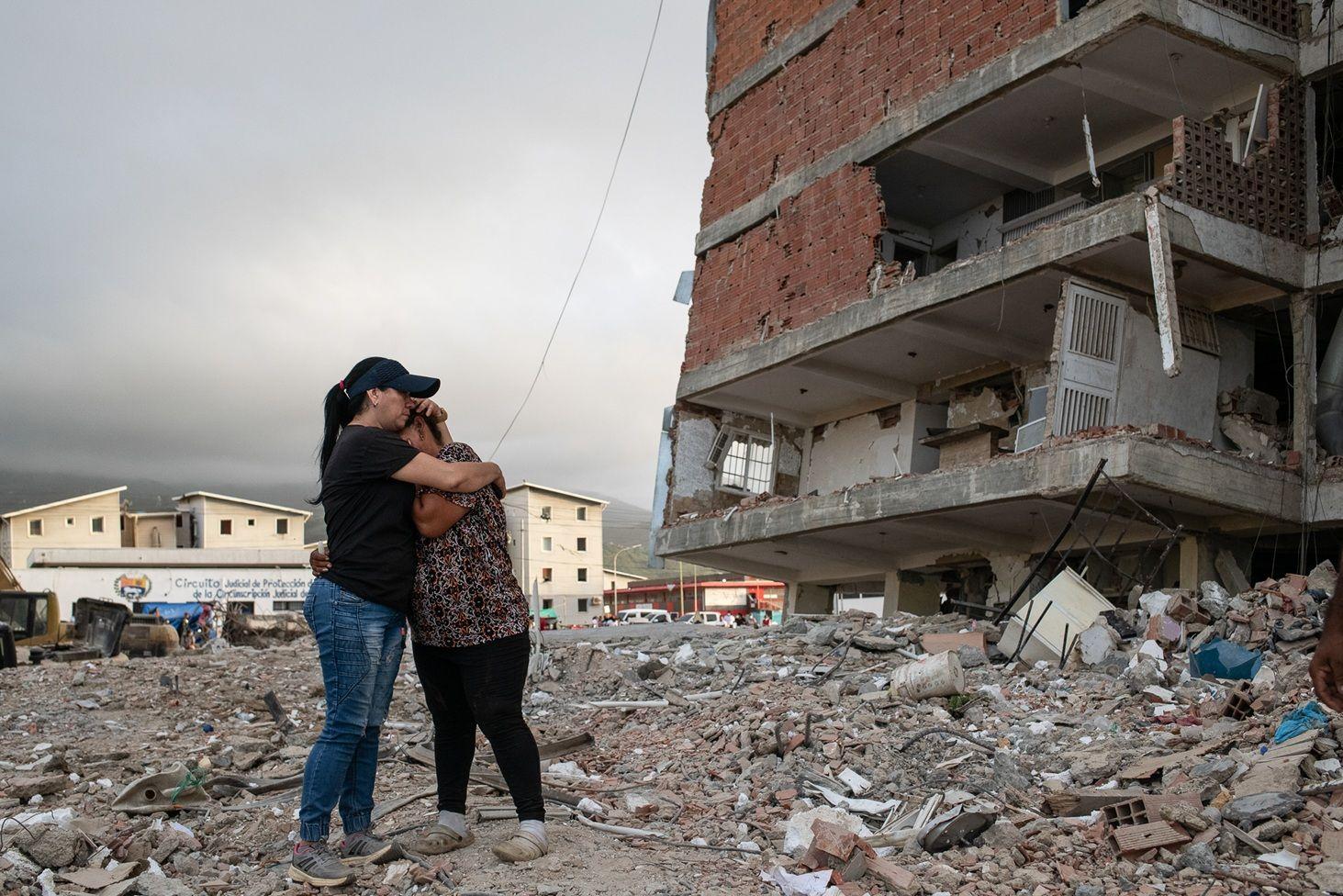 Dos mujeres paradas se abrazan en medio de los escombros y frente a un edificio gravemente afectado.
