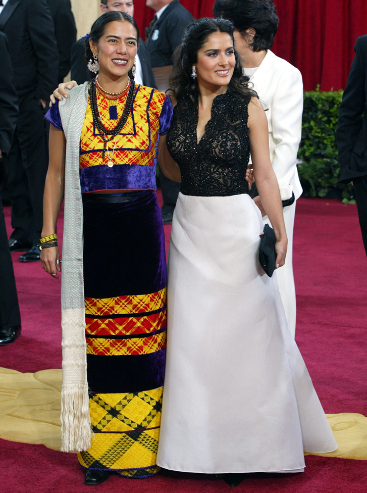 Lila Downs y Salma Hayek posan sonrientes frente a las cámaras en la alfombra roja. Downs lleva un vestido tradicional de México de colores amarillo, azul, negro y rojo. Hayek lleva un vestido largo de colores blanco y negro