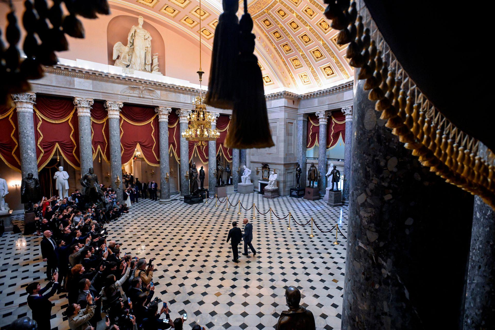 Širok kadar prikazuje kralja kako ga vode kroz hodnik Kapitola, dok su reporteri sa strane.
