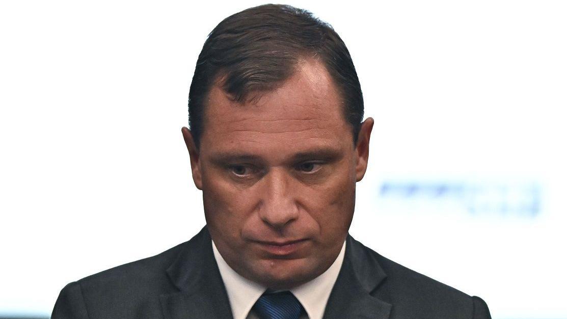 Mauro Cid com olhar consternado, olhando levemente para baixo em frente a fundo branco