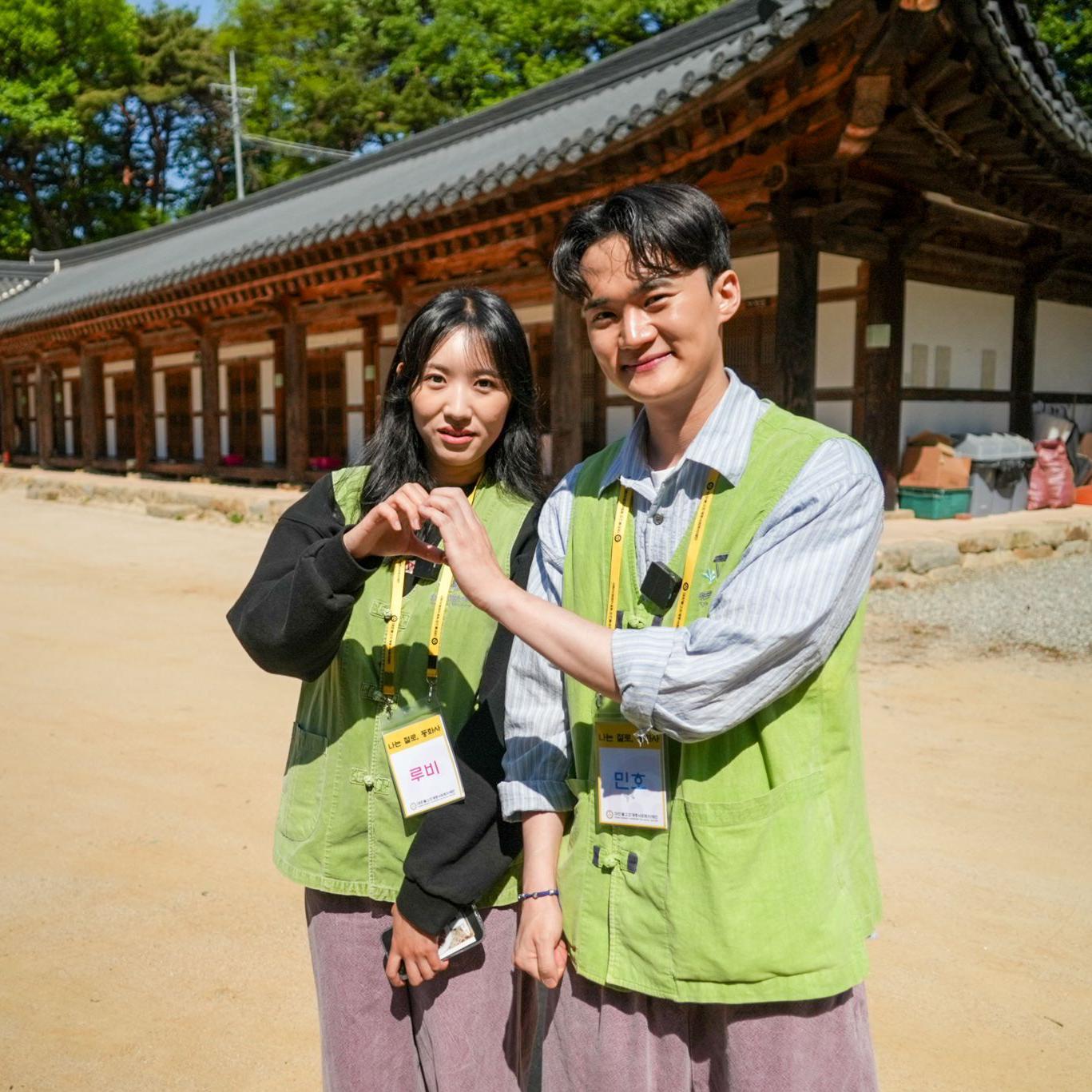 루비와 민호가 연두색 조끼와 행사 명찰을 착용한 채 마당에 서서 손을 맞잡아 하트 모양을 만들고 있다.