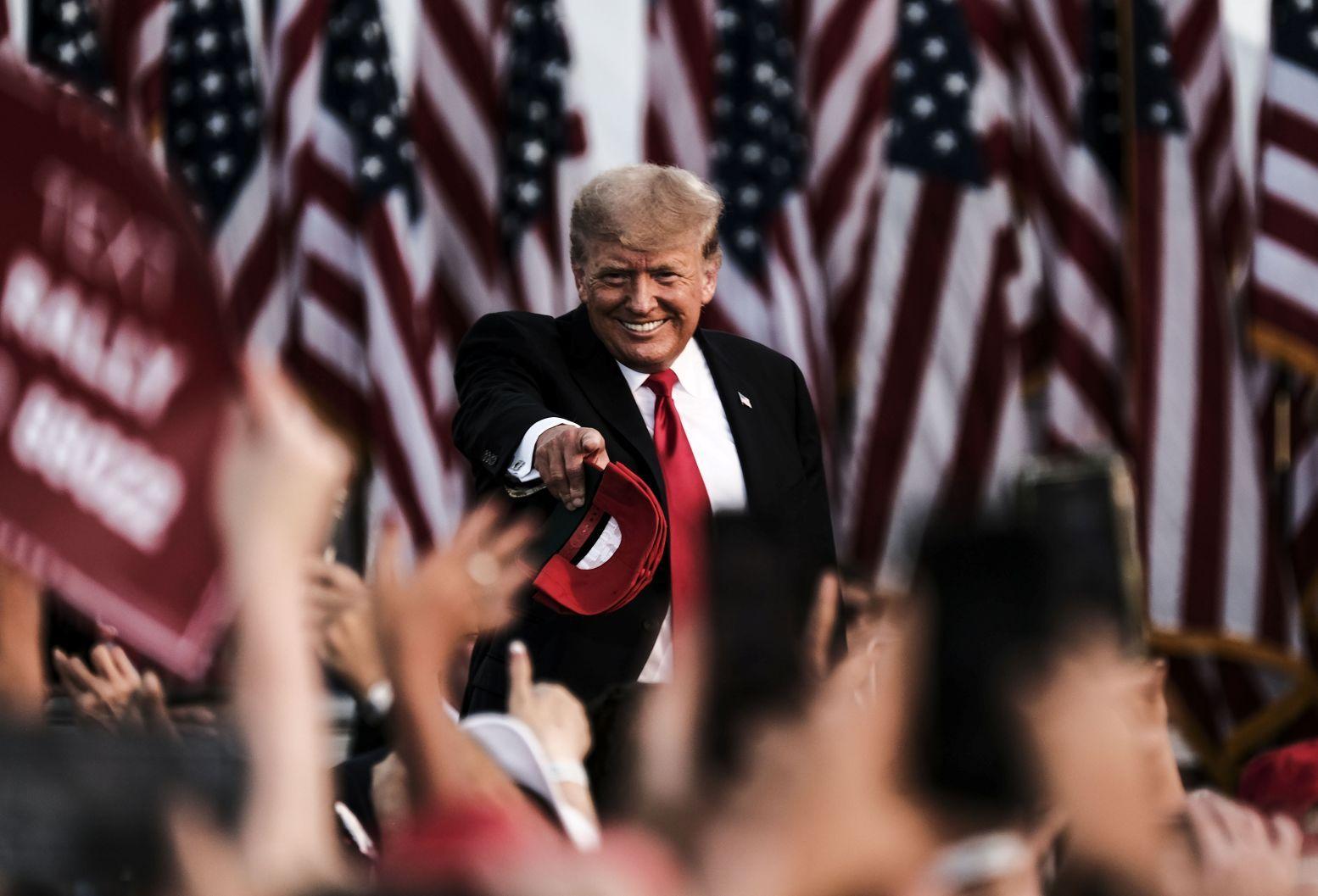 Donald Trump apontando para multidão em comício em 2021, com várias bandeiras dos EUA atrás dele