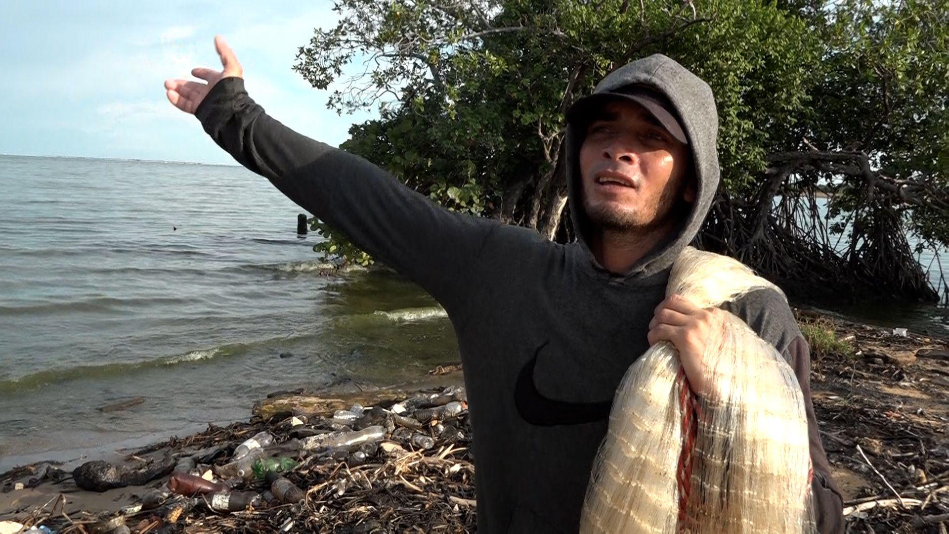 Un hombre vistiendo una sudadera gris con capucha y una gorra negra, mientras sostiene con su mano izquierda una red de pesca sobre su hombro izquierdo, y su otra mano apunta al mar.