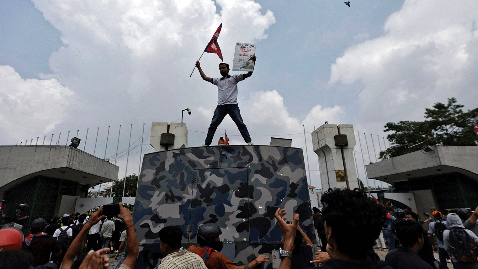Manifestantes na porta do parlamento. Um homem está em cima de uma estrutura segurando uma bandeira