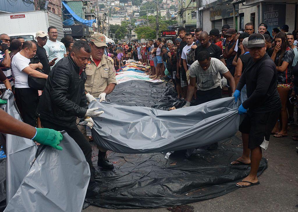 Policiais carregam um corpo no Rio de Janeiro. Atrás deles há uma fila de corpos no chão.