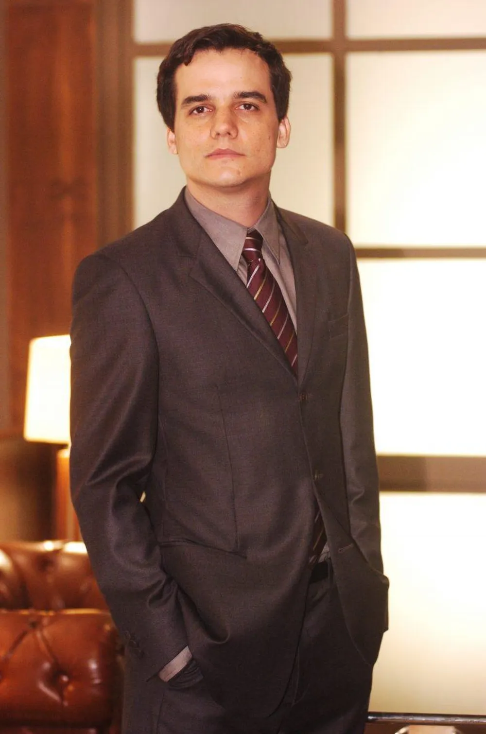 Wagner Moura aparece como Olavinho em cena da novela Para&iacute;so Tropical. Ele veste terno escuro e gravata, est&aacute; de p&eacute; com as m&atilde;os nos bolsos e olha com semblante s&eacute;rio para a c&acirc;mera.