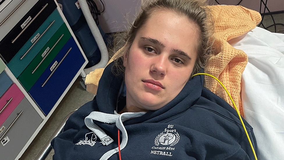 Phoebe Tesoriere deitada em uma cama de hospital, com agasalho azul