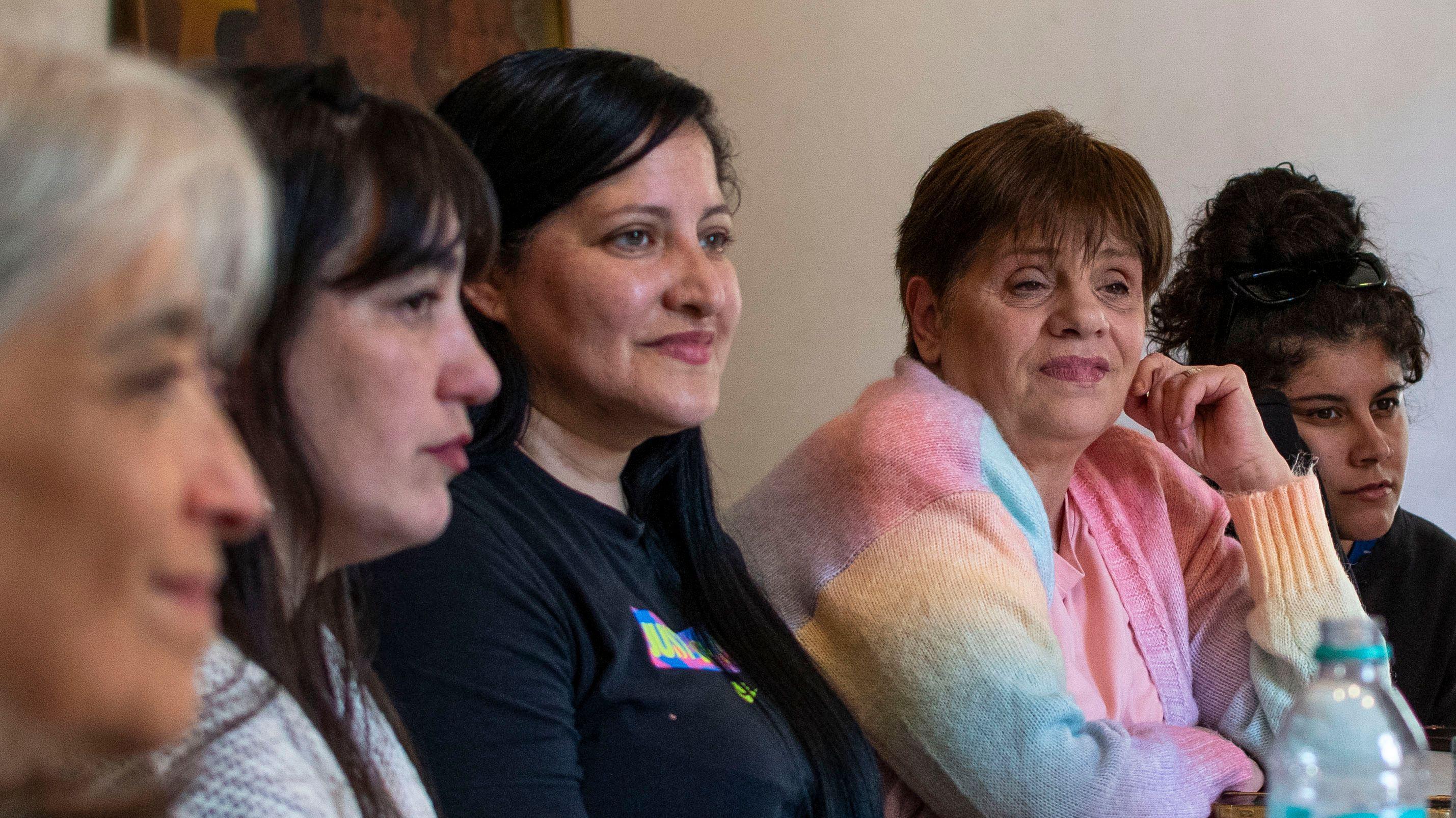 Cinco mujeres de diferentes edades sentadas a lo largo de una mesa mirando hacia una misma dirección. Andrea, la cuarta de izquierda a derecha, luce un saco de colores pastel y se lleva una mano a la cara.