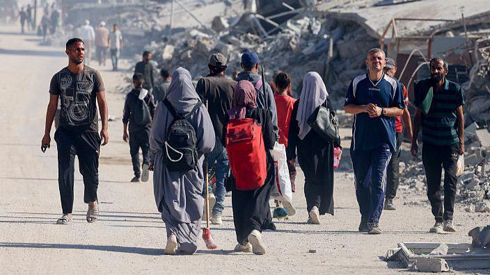 Varias personas caminando por Gaza, la mayoría lejos de la cámara, con enormes pilas de escombros en el fondo.
