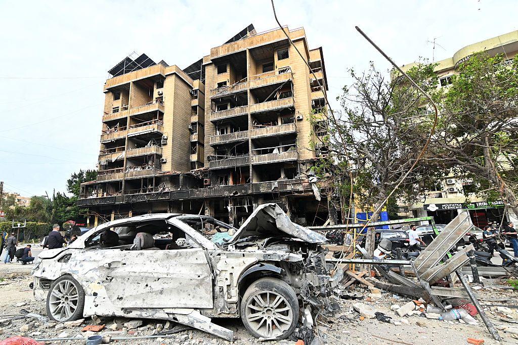 Carro branco seriamente danificado em frente a um edif&iacute;cio fortemente atingido, rodeado por destro&ccedil;os