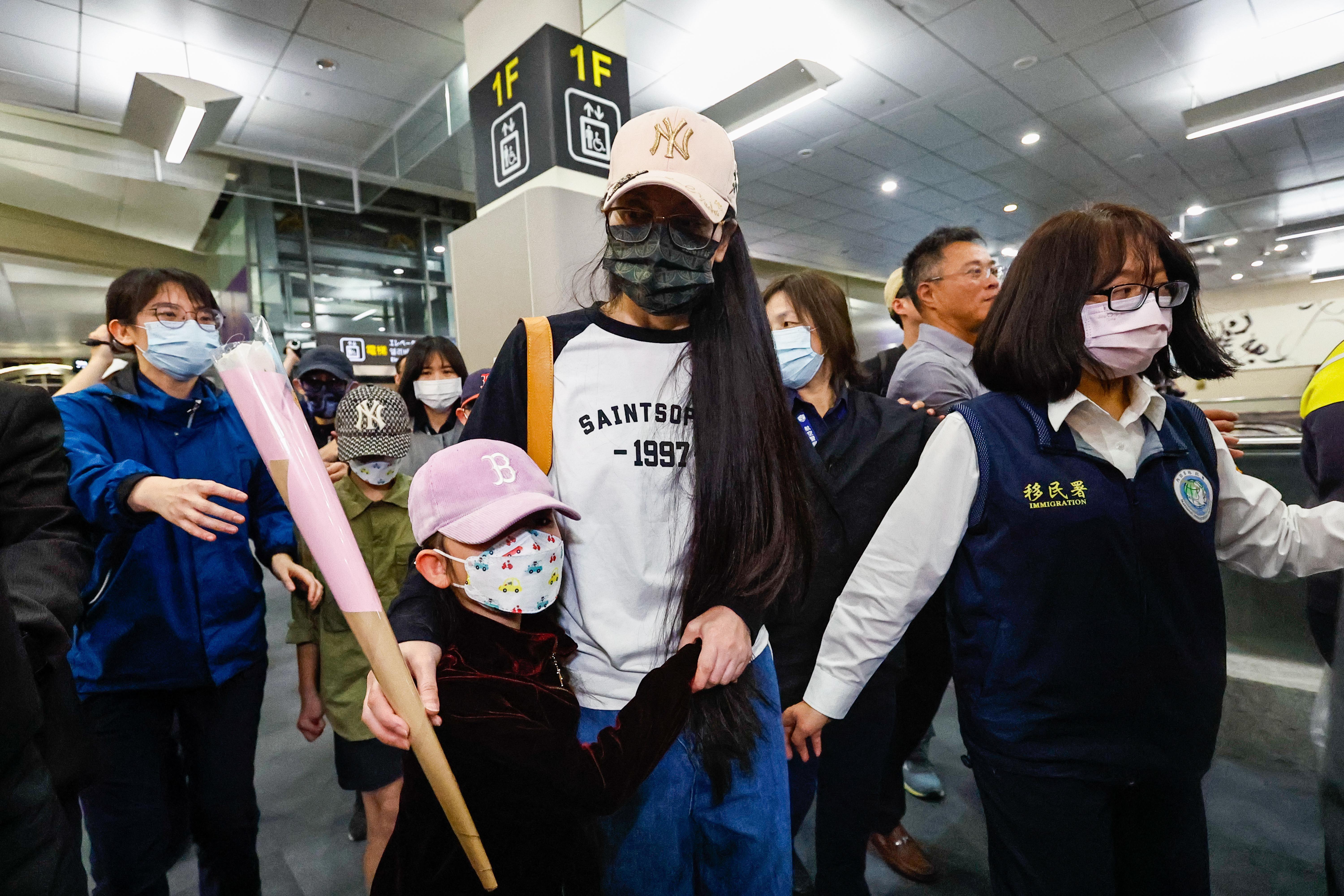 大陸配偶網紅「亞亞」在台灣的機場，她戴上口罩和米黃色的鴨舌帽，擁著一名兒童，旁邊有身穿寫有「移民署」的人士陪同。