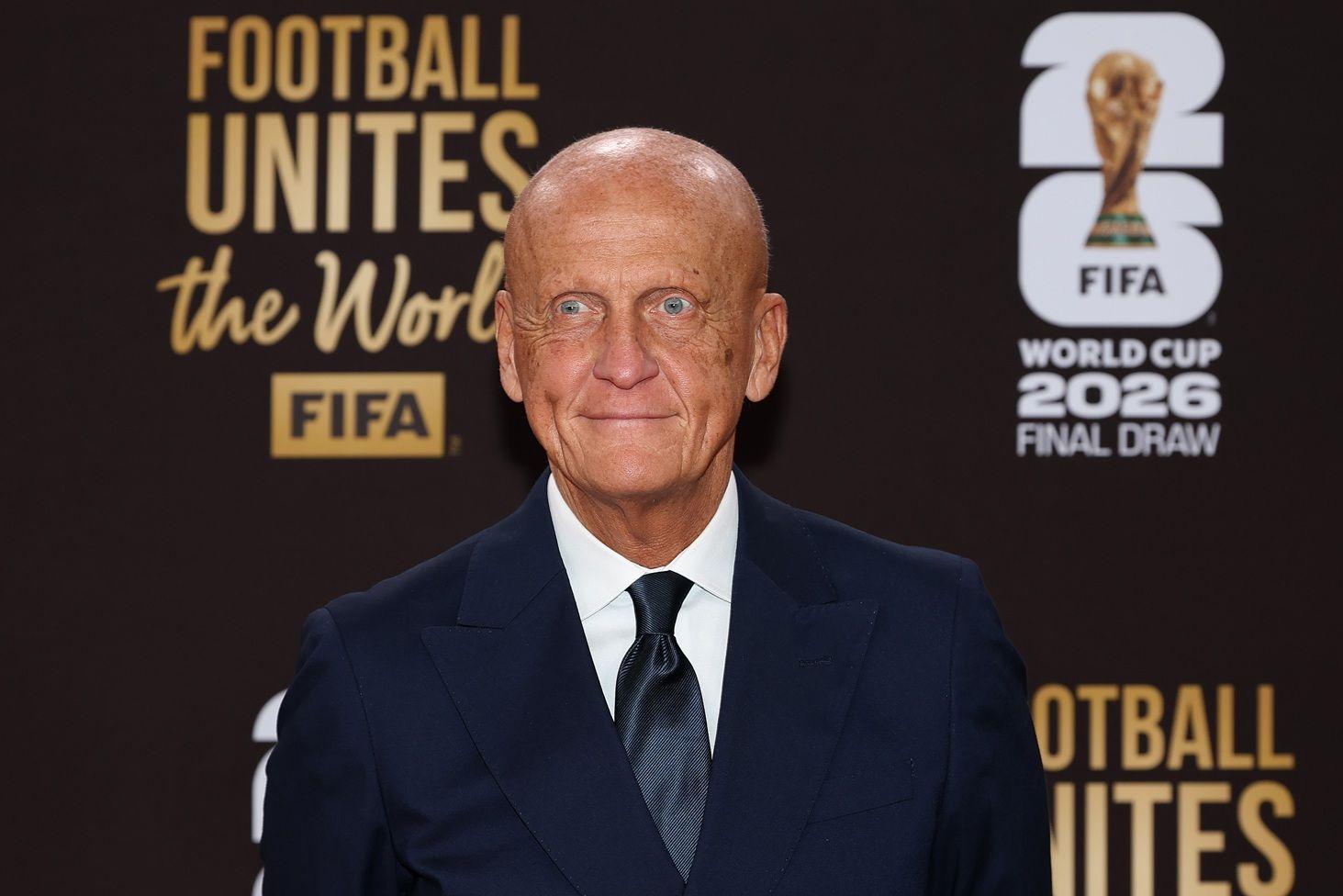 Pierluigi Collina, vestido de traje negro y corbata negra con una camisa blanca, está sentado en el Centro John F. Kennedy. Atrás tiene una pancarta que dice "Football Unites The  World. FIFA World Cup 2026 Final Draw" y tiene una foto de la Copa del mundo