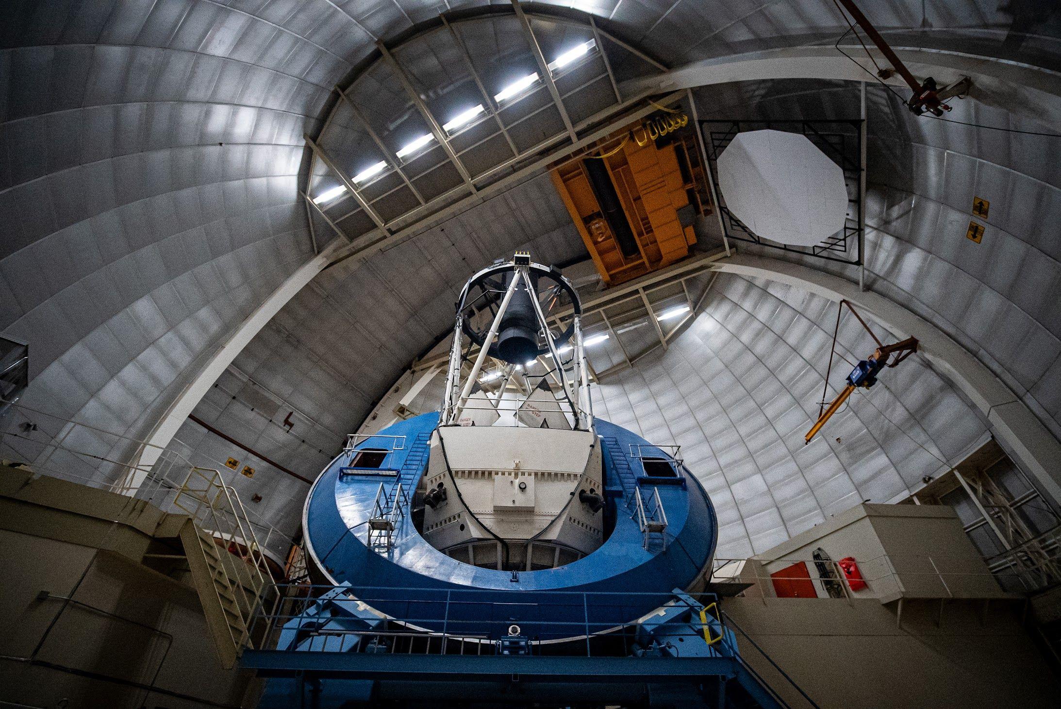 The photo shows the inside of a huge metallic dome, with curved silver panels arching high overhead like the inside of a giant steel shell.In the centre stands a massive blue and white telescope mount, shaped like a thick ring with a tall open framework above it that holds the main mirror and instruments.Walkways, railings and ladders cling to the structure, and an orange overhead crane hangs near the ceiling, giving the scene an industrial, cathedral‑like feel.