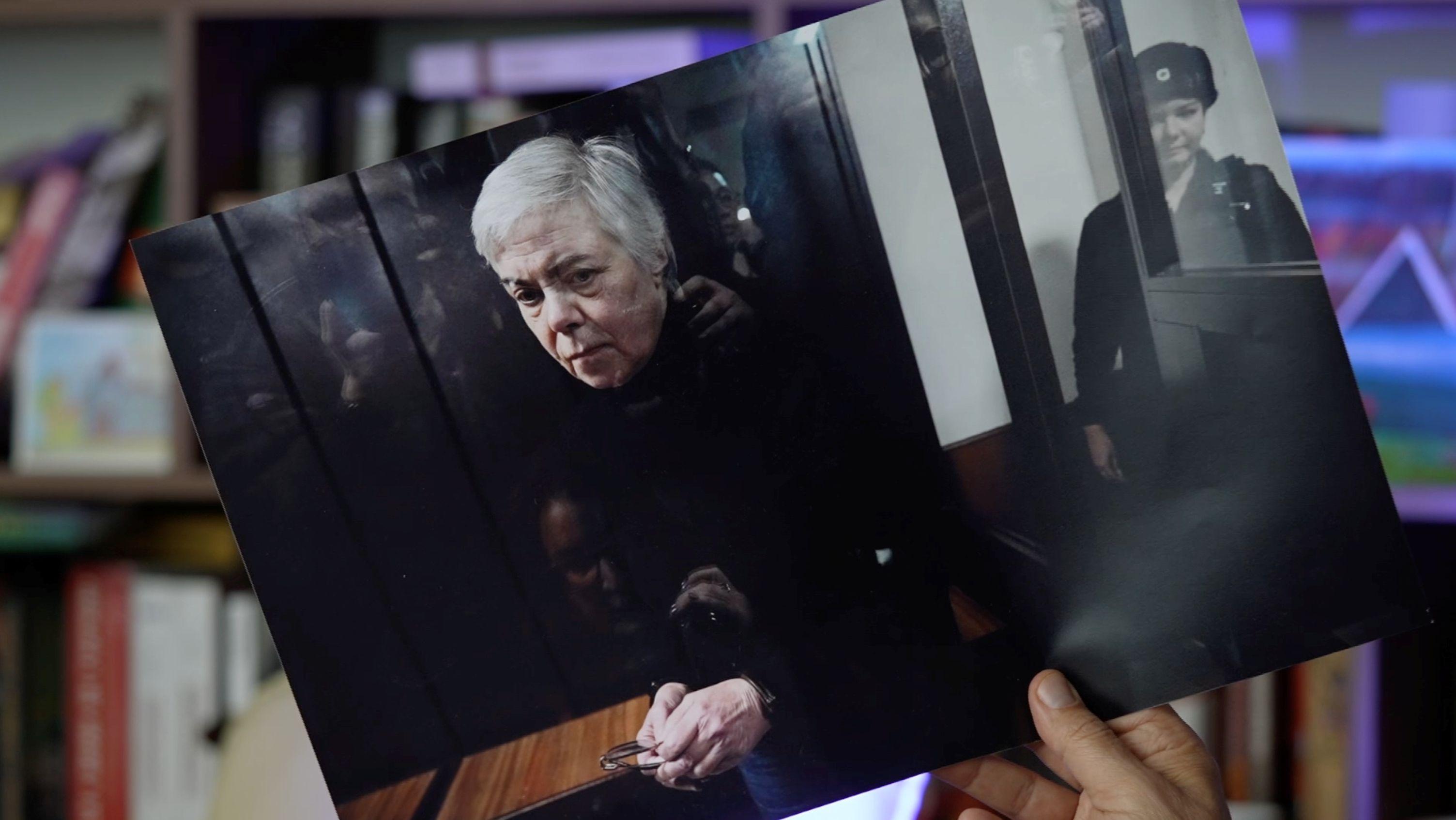 Uma m&atilde;o segura uma foto impressa de Nadezhda Buyanova, pediatra de 68 anos nascida na Ucr&acirc;nia, no banco dos r&eacute;us e sob escolta durante o julgamento