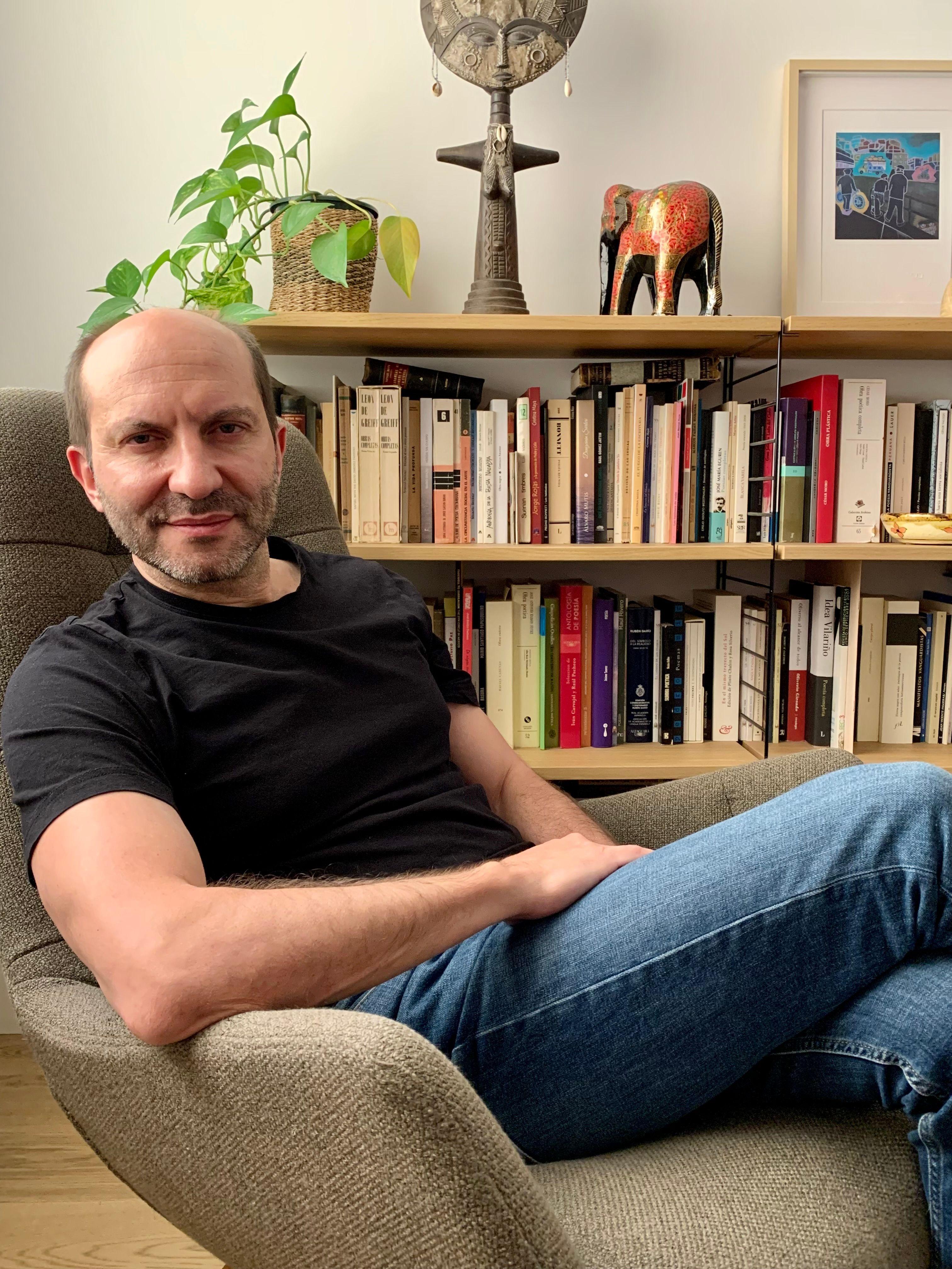 Carlos Granés, está sentado en una silla con jeans y camiseta negra. Detrás tiene una biblioteca con libros. 