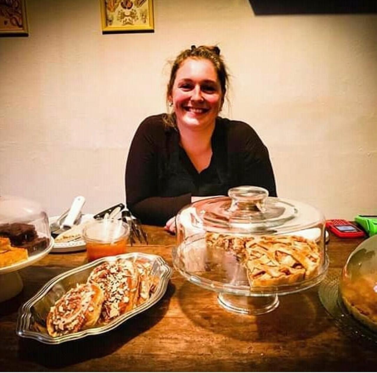 A imagem mostra uma mulher sorrindo atr&aacute;s do balc&atilde;o, cercada por doces e tortas expostos em pratos e redomas de vidro. Vestindo roupas escuras e com os cabelos presos, ela apoia os bra&ccedil;os sobre a bancada de madeira.
