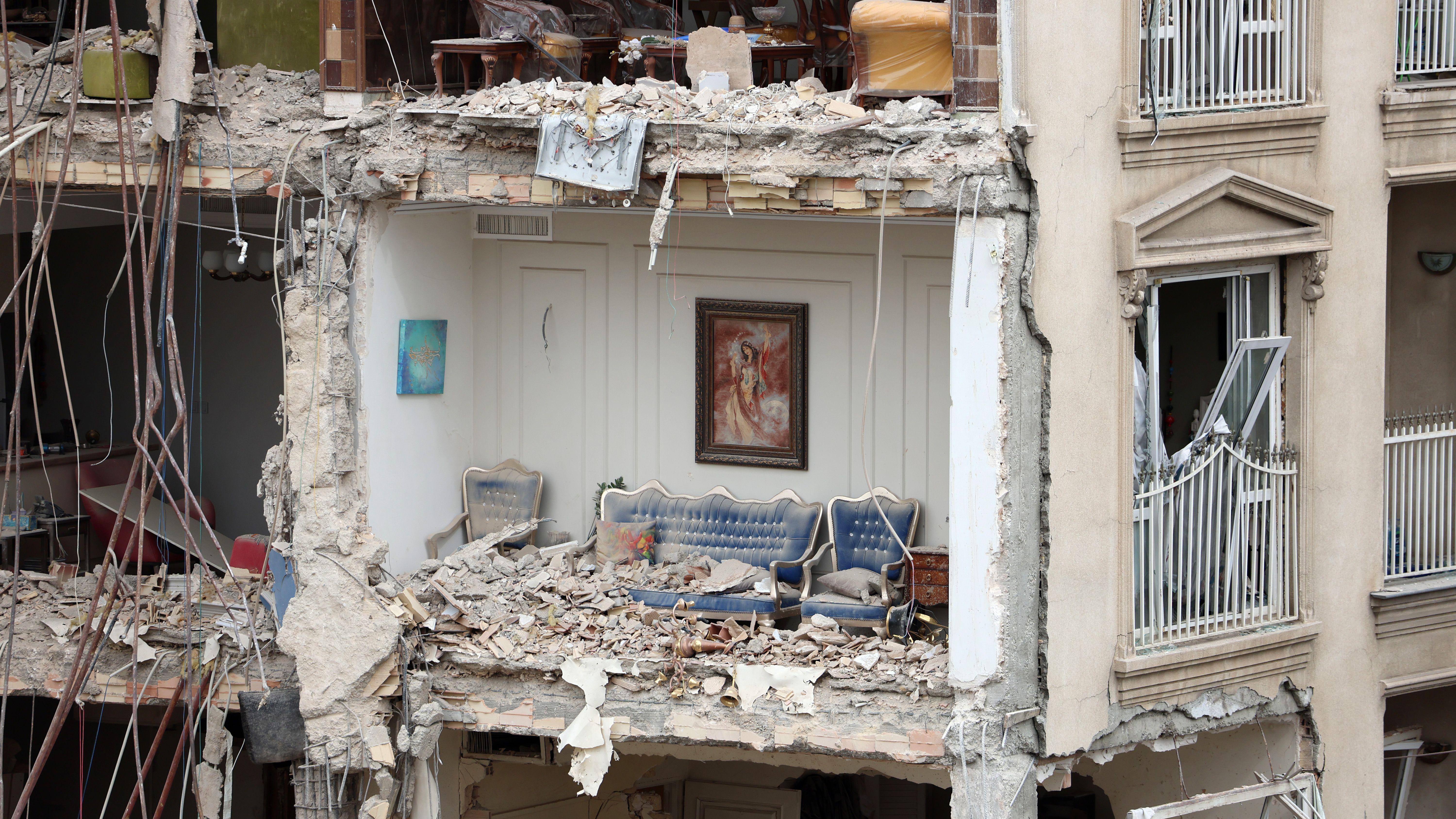 Bloco de apartamentos residenciais parcialmente destru&iacute;do em Teer&atilde;, sem a parede externa e com um sof&aacute; azul em um c&ocirc;modo repleto de escombros e um quadro ainda pendurado na parede