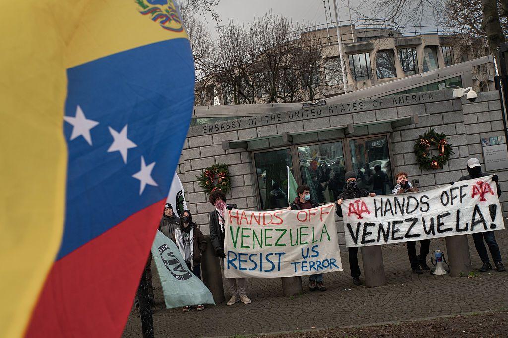 En primer plano, bandera venezolana, de fondo, personas manifestándose con pancartas donde se puede leer en inglés "Hands off Venezuela" - manos fuera de Venezuela. 