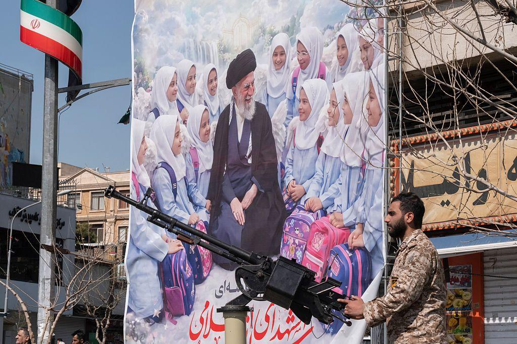 Membro armado da Guarda Revolucion&aacute;ria Isl&acirc;mica do Ir&atilde;, de guarda atr&aacute;s de uma arma, em frente a um banner ilustrando o ex-l&iacute;der supremo iraniano, o aiatol&aacute; Ali Khamenei e meninas de uma escola