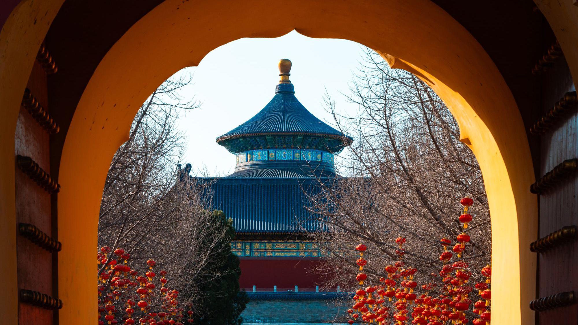 Una vista a través de un portal histórico, contemplando los ornamentados edificios de un templo en tonos azules, rojos y dorados, con numerosas linternas chinas colgando de los árboles desnudos en primer plano.