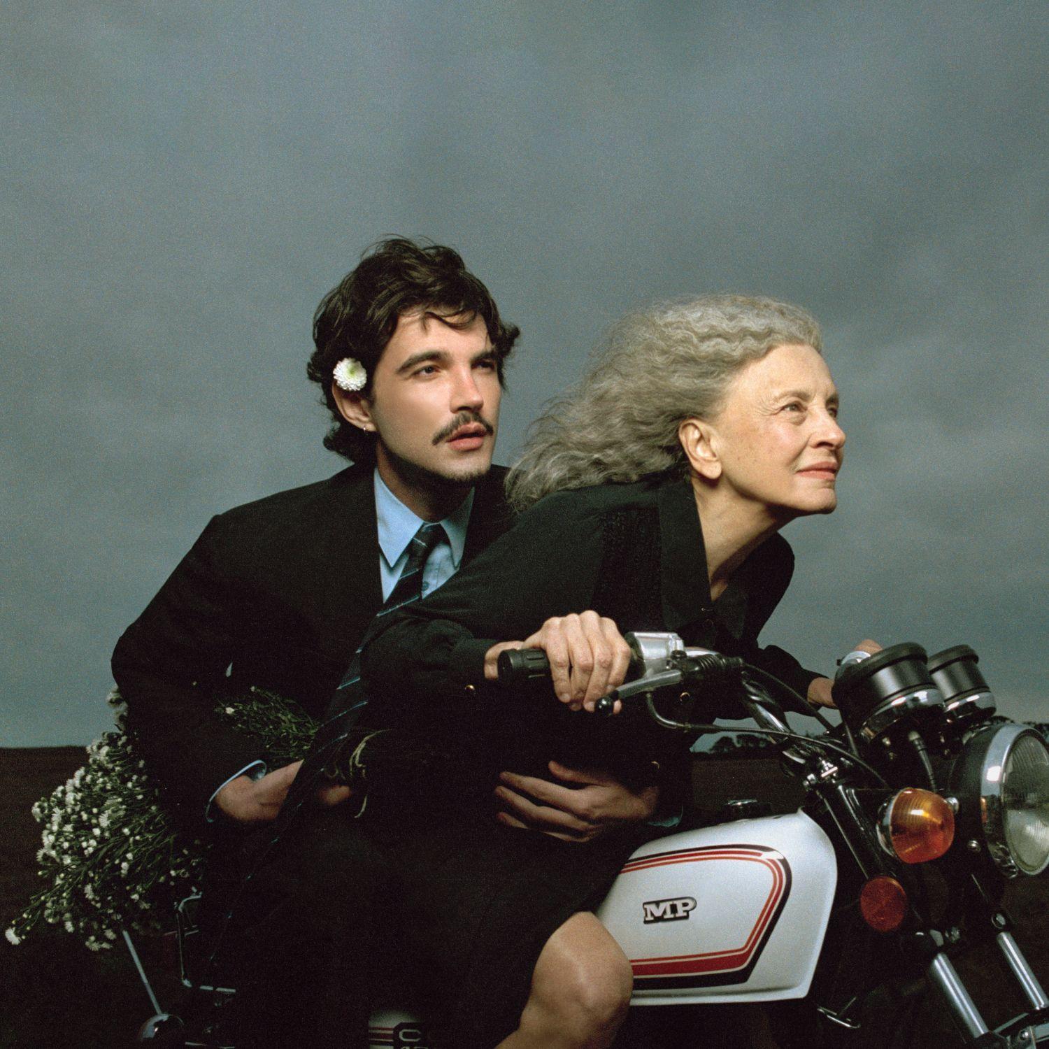 Um homem jovem (Jão) e uma mulher idosa (Marisa Sabino dos Santos) estão sobre uma motocicleta branca, ambos vestidos de preto e olhando para a frente. O homem usa terno, camisa azul, gravata e uma flor branca atrás da orelha, enquanto a mulher, de cabelos grisalhos, segura o guidão da moto. Na parte traseira do veículo há um buquê de flores brancas, e o fundo é formado por um céu cinzento.