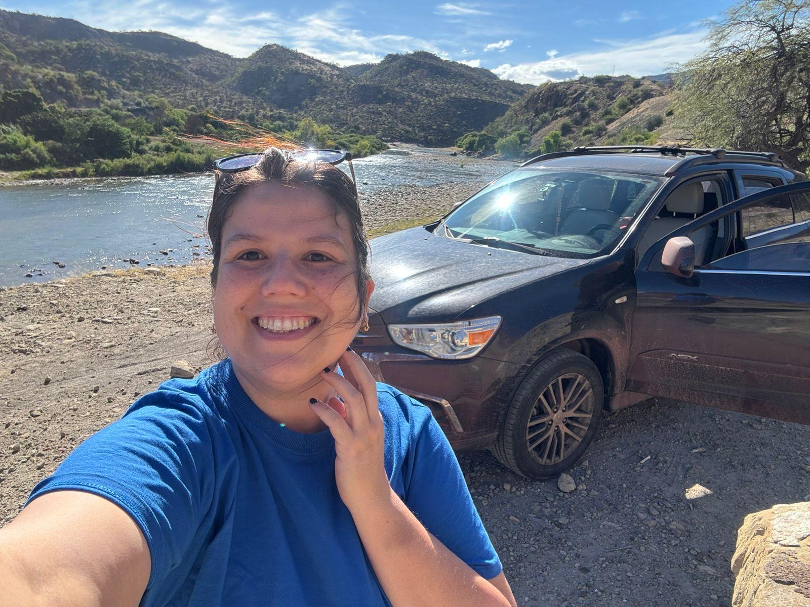 Fabiane fazendo uma selfie com um lago ao fundo. Atr&aacute;s dela est&aacute; o carro em que ela viajava