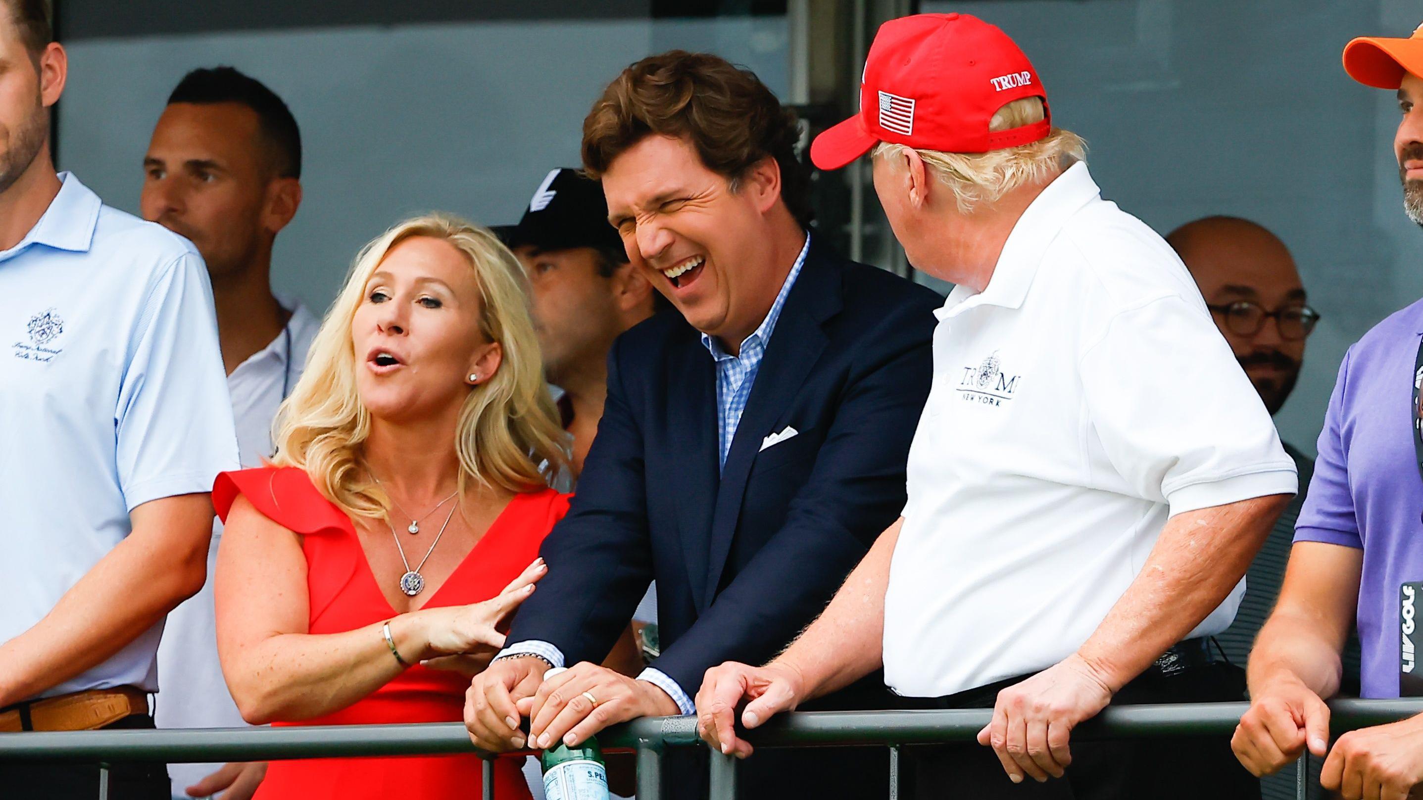 Marjorie Taylor Greene de vestido vermelho, ao lado de Tucker Carlson de terno, ouvindo Donald Trump de costas para a c&acirc;mera