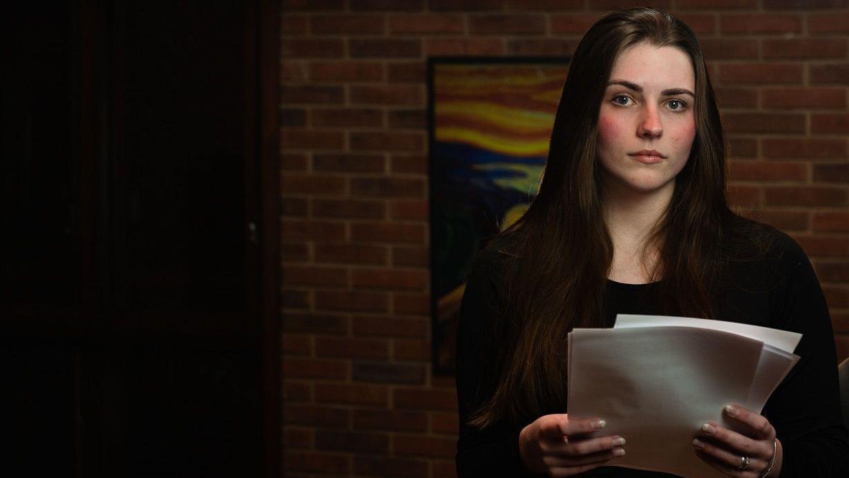 Michele olhando sério para cama, em sala de casa, segurando papelada