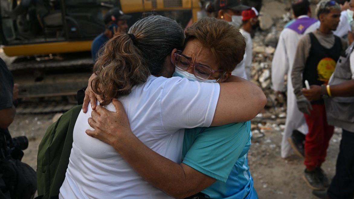 Dos mujeres se abrazan, una lleva una mascarilla y lentes. Están en una zona devastada. Detrás de ellas hay una maquinaría y varias personas, entre ellas un sacerdote
