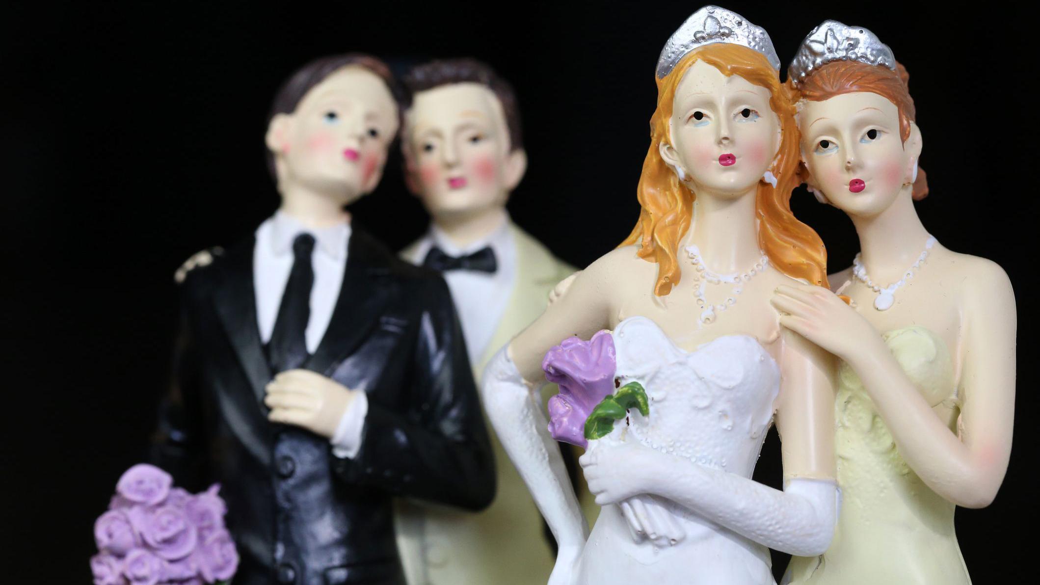 Imagem de 4 bonecos, dois vestidos de noivos e dois vestidos de noivas, como se fossem decoração de bolo de casamento. 