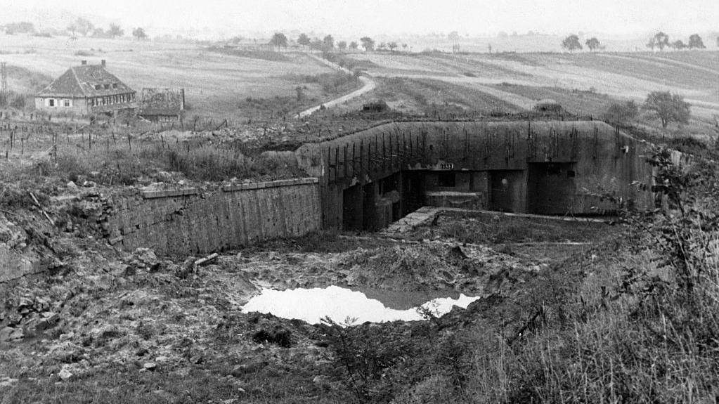 Bunker destruído da Segunda Guerra Mundial, em um campo aberto com um casarão ao fundo
