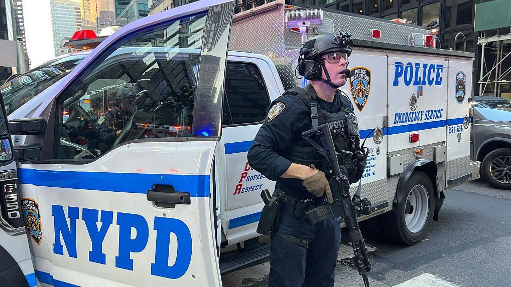 Un policía armado frente a un vehículo policial monta guardia atento en una avenida de Nueva York
