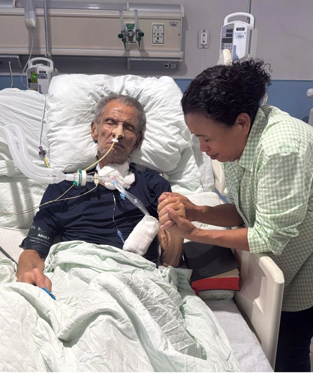 Brooklyn Rivera está acostado en una cama de hospital con un tubo conectado a su nariz. Nancy Elizabeth Henríquez, quien está parada a su lado, le agarra una mano con sus dos manos mientras cierra sus ojos