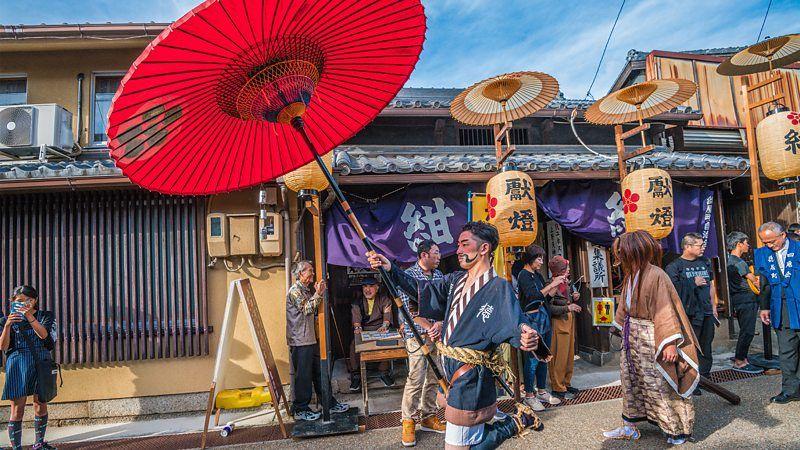 Desfile pelas ruas no Japão, com um homem empunhando um guarda-chuva vermelho à frente
