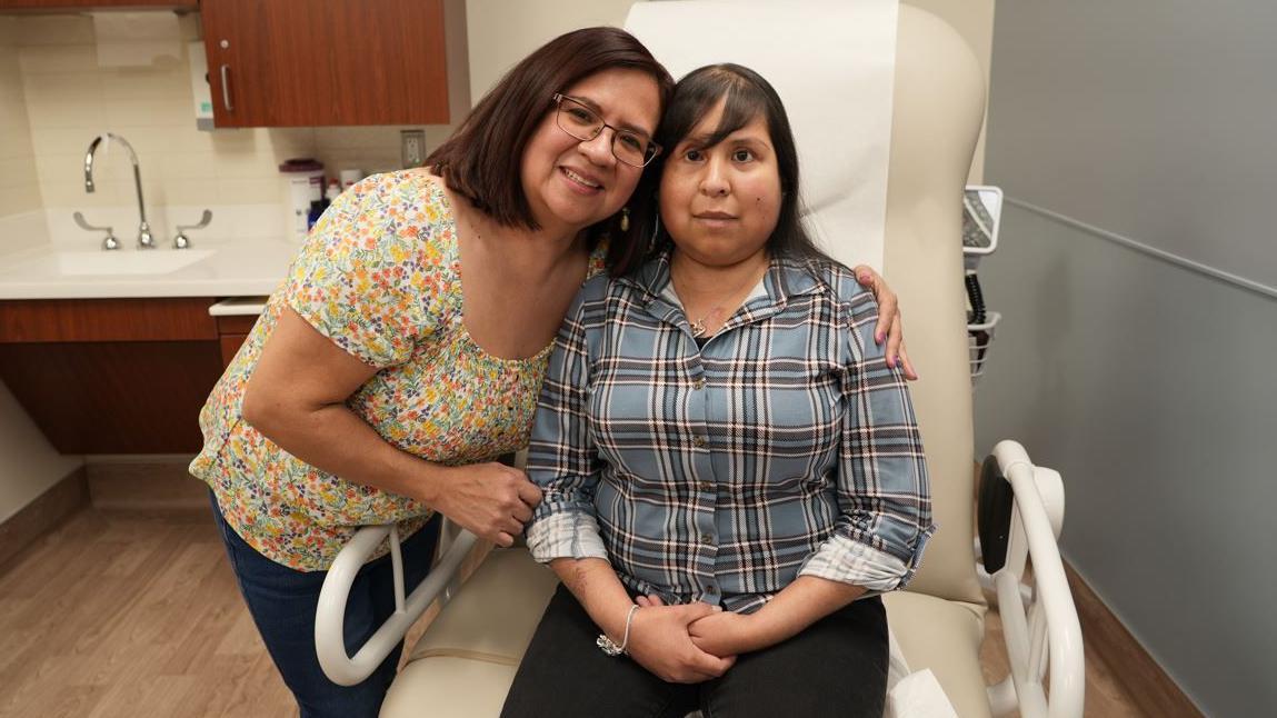 Jessica López sentada en una silla del hospital, vestida con pantalones negros y una camisa, recibiendo un cálido abrazo de su madre.