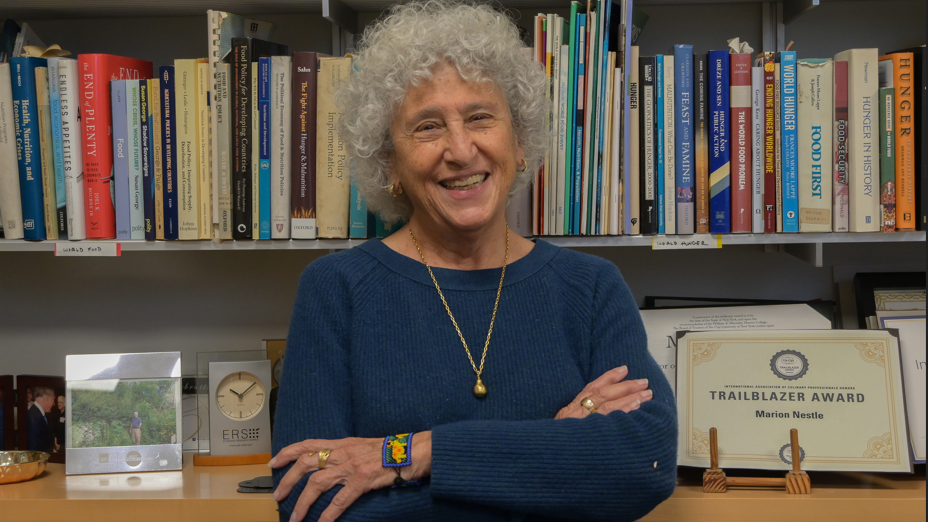 Nestle sorrindo, de braços cruzados, em frente a prateleiras com livros e prêmios