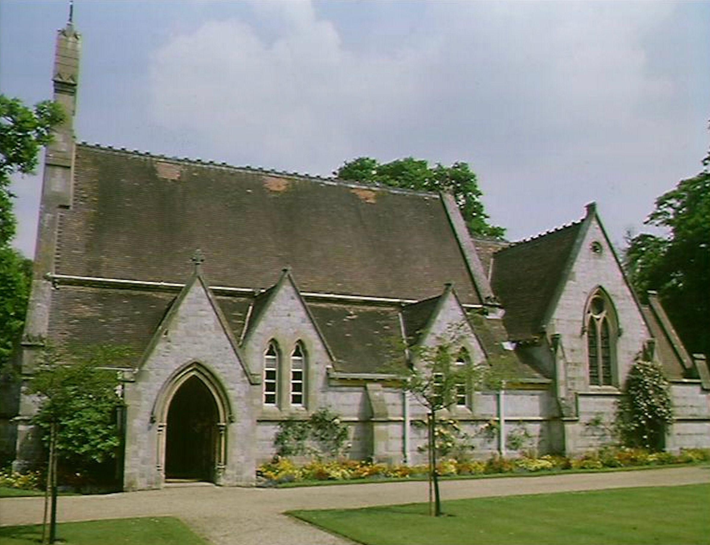 A chapel with a spire, pitched roof and arched entrance. A path and neat lawns are in front of it with small trees growing on the lawns.