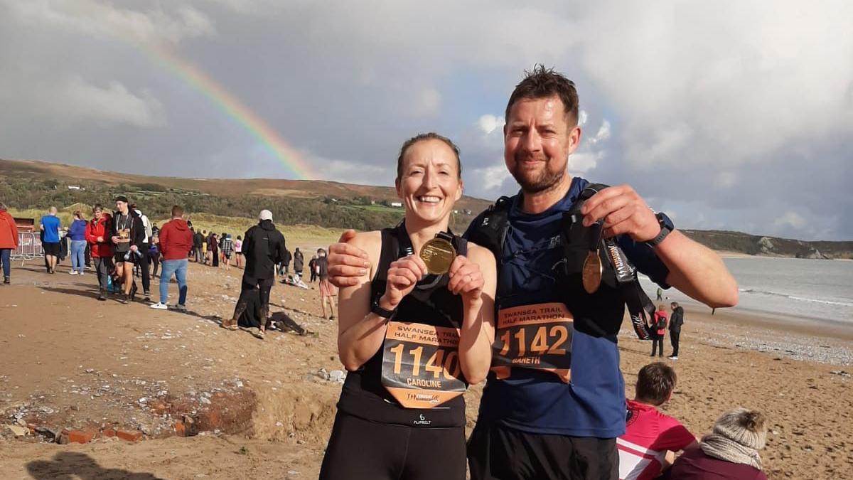 Gareth e Caroline, com roupas de corrida, na praia de Swansea (Pa&iacute;s de Gales). Eles est&atilde;o sorrindo e segurando medalhas de corrida