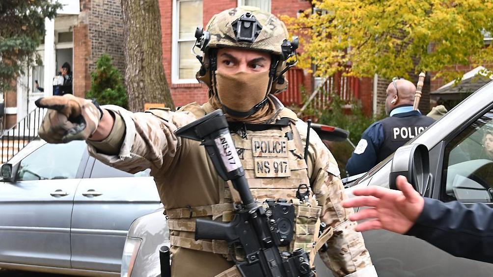 Un agente del Servicio de Inmigración y Aduanas de Estados Unidos, ICE, con un uniforme de campaña, casco, un pasamontañas que la cubre la cara parcialmente y un rifle de asalto señala con el dedo hacia un lado, como dando órdenes