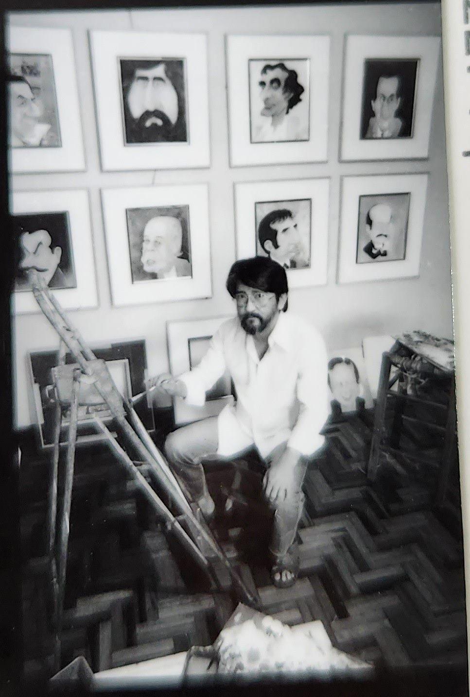 A imagem em preto e branco mostra Cl&aacute;udio Seto, um homem barbudo e de &oacute;culos, agachado em um ateli&ecirc; ou sala de trabalho, cercado por quadros com caricaturas pendurados na parede. &Agrave; frente dele h&aacute; um cavalete de madeira, enquanto outros retratos apoiados no ch&atilde;o e m&oacute;veis aparecem ao redor. O ambiente tem piso de madeira em padr&atilde;o geom&eacute;trico e aspecto de arquivo fotogr&aacute;fico antigo.