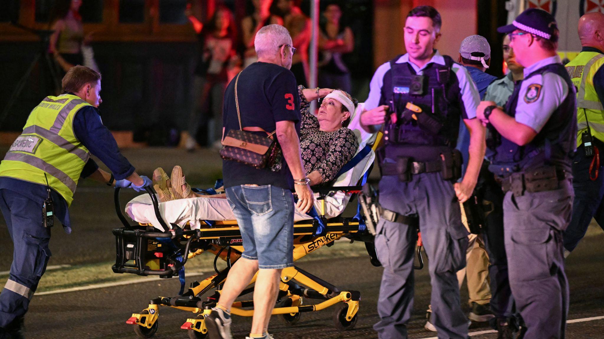 悉尼邦迪海灘救護員用擔架將一名清醒的年長女傷者（中）送走，警員（右）在旁交談（14/12/2025）